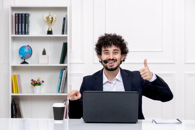 Servizio clienti bell'uomo riccio in tuta da ufficio con computer e auricolare felice per un caffè