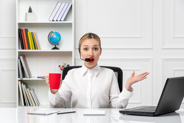 Servizio clienti bella ragazza bionda in camicia bianca con laptop e auricolare confuso con tazza rossa