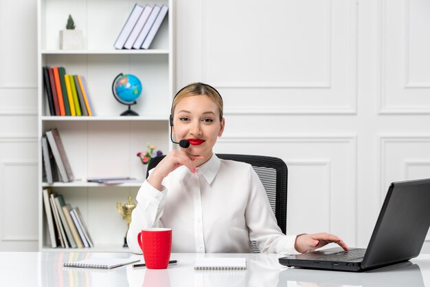 Servizio clienti bella ragazza bionda in camicia bianca con laptop e auricolare sorridente con tazza rossa