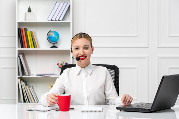 Servizio clienti bella ragazza bionda in camicia bianca con laptop e auricolare sorridente e tenendo la tazza