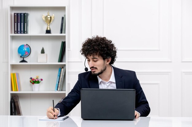 Servizio clienti carino ragazzo bruna in abito da ufficio con computer e auricolare scrivendo note