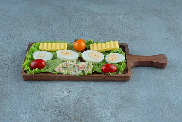 Set colazione su un vassoio in legno con verdure, uova sode, burro e insalata, su fondo in marmo. Foto di alta qualità