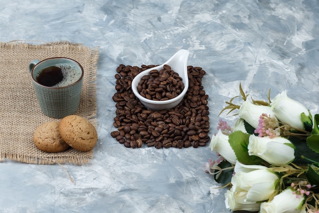 Set di biscotti, tazza di caffè, fiori e chicchi di caffè in una brocca di porcellana bianca su uno sfondo di marmo blu. avvicinamento.