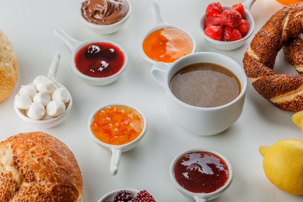 Set di marmellate, lamponi, zucchero, cioccolato in tazze, bagel turco, pane, limone e una tazza di caffè su una superficie bianca