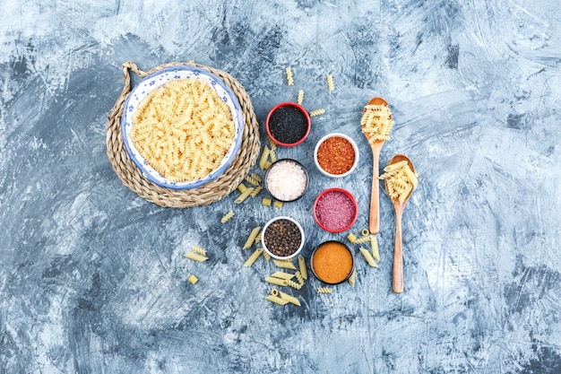 Set di spezie, cucchiai di legno e pasta fusilli in una ciotola su intonaco grigio e sfondo tovaglietta di vimini. vista dall'alto.