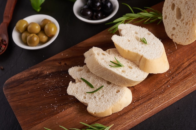 Set di spuntini mediterranei. Olive, olio, erbe aromatiche e ciabatta a fette su una tavola di legno su ardesia nera tavola di pietra sulla superficie scura, vista dall'alto. Lay piatto.