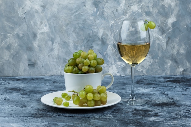 Set di un bicchiere di vino e uva verde in tazza bianca e piatto su uno sfondo di gesso grungy. vista laterale.