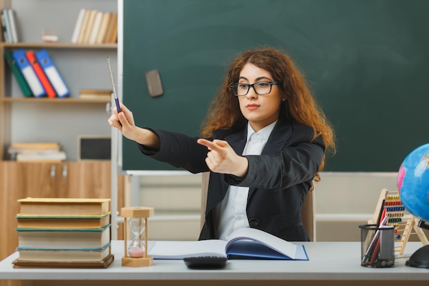 severa giovane insegnante femminile che indossa occhiali punta a lato con il puntatore seduto alla scrivania con strumenti scolastici in classe