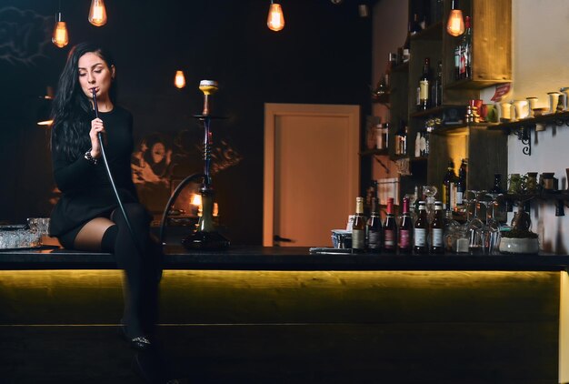 Sexy ragazza bruna in seducenti abiti neri fuma un narghilè mentre è seduto sul bancone in una discoteca.