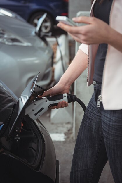 Sezione centrale della donna che utilizza il telefono cellulare durante la ricarica dell'auto elettrica