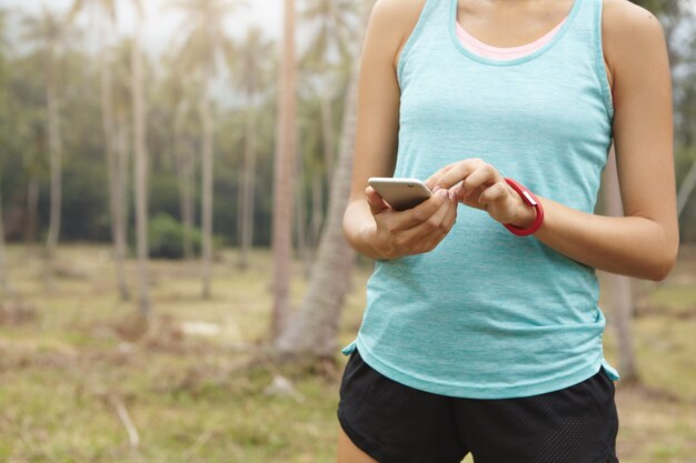 Sezione mediana del jogger femminile in abbigliamento sportivo che tiene il cellulare, utilizzando il fitness tracker dell'app per monitorare i progressi nella perdita di peso durante l'allenamento cardio.