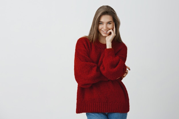 Sfacciata e civettuola attraente fidanzata in elegante maglione sciolto rosso invernale sorridente ingannevole e sensuale dalla soddisfazione e dalla gioia toccando lo sguardo delle labbra assicurato e fiducioso