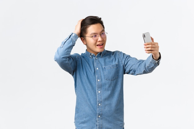 Sfacciato uomo asiatico fiducioso con gli occhiali e l'apparecchio che si sente impertinente, scatta selfie, si spazzola i capelli con la mano, posa per una foto, usa l'app filtro sul cellulare, il blogger fa post su internet.