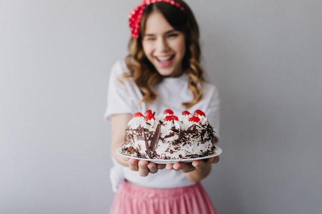 Sfocatura ritratto di jocund donna caucasica in posa con torta di compleanno e sorridente Indoor shot di ridere modello femminile con torta cremosa in primo piano