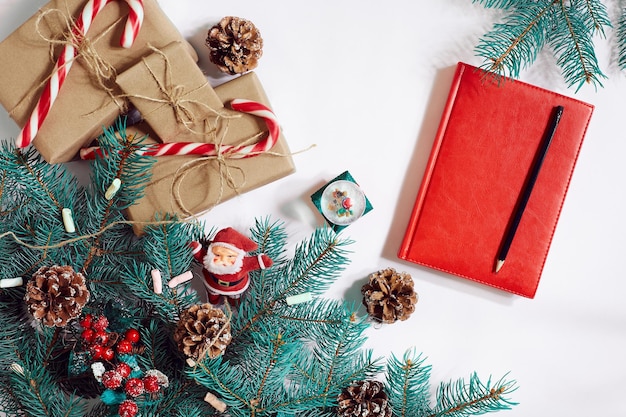 Sfondo di Natale o Capodanno: albero di pelliccia, rami, regali, decorazioni, taccuino rosso con penna su sfondo bianco