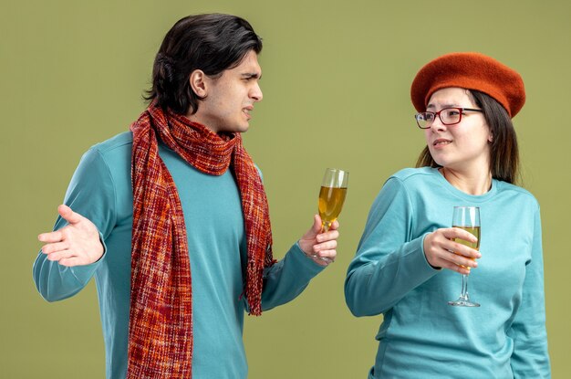 Sgradevole giovane coppia il giorno di san valentino ragazzo che indossa una sciarpa ragazza che indossa un cappello con in mano un bicchiere di champagne guardando l'altro isolato su sfondo verde oliva