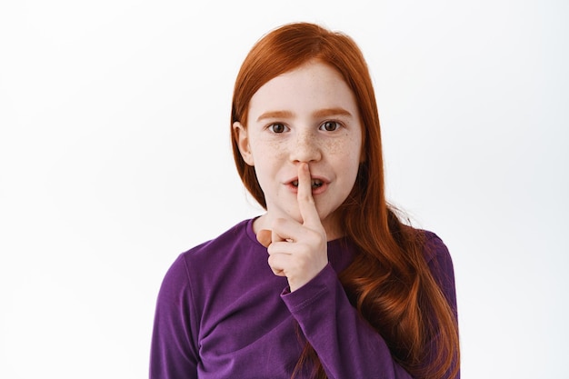 Shh questo è segreto. Carina bambina rossa, bambino che zittisce, mostra taci gesto tabù, dito sulle labbra, silenzio, in piedi su sfondo bianco