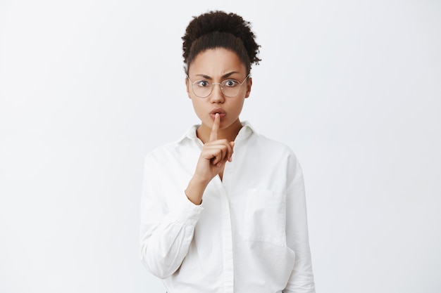 Shh, tranquillo, silenzio in classe. Insegnante femminile dalla pelle scura rigorosa e dall'aspetto serio in camicia da colletto bianco e occhiali che fa un gesto di silenzio con il dito indice sulla bocca, chiedendo di mantenere la voce bassa