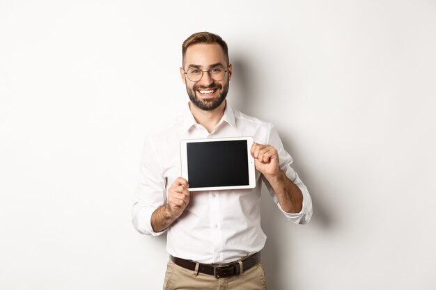 Shopping e tecnologia. Uomo bello che mostra lo schermo della tavoletta digitale, con gli occhiali con camicia bianca, sfondo per studio.