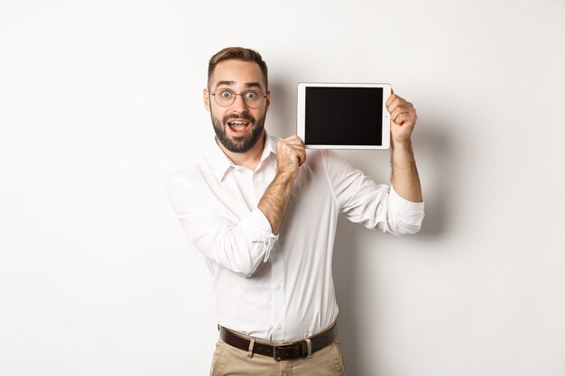 Shopping e tecnologia. Uomo bello che mostra lo schermo della tavoletta digitale, con gli occhiali con camicia bianca, sfondo per studio.