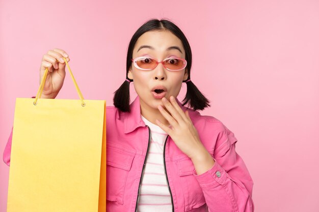 Shopping Elegante ragazza asiatica in occhiali da sole che mostra la borsa dal negozio e sorridente consigliando promo di vendita in negozio in piedi su sfondo rosa