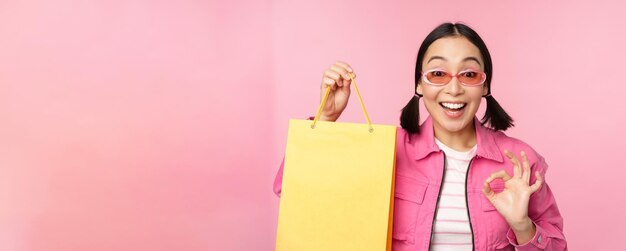 Shopping Elegante ragazza asiatica in occhiali da sole che mostra la borsa dal negozio e sorridente consigliando promo di vendita in negozio in piedi su sfondo rosa