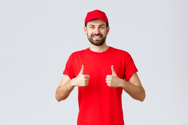 Shopping online, consegna durante la quarantena e concetto da asporto. Corriere allegro in berretto e t-shirt uniforme rossa, consiglia di effettuare ordini, pollice in su in approvazione, sfondo grigio