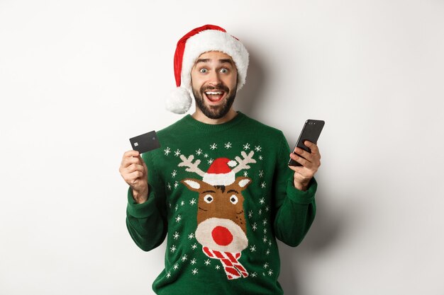 Shopping online e concetto di vacanze invernali. Uomo sorpreso con cappello da Babbo Natale, con in mano un telefono cellulare e una carta di credito, in piedi con un maglione su sfondo bianco