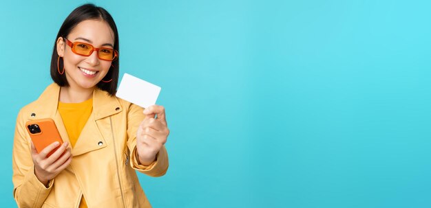 Shopping online elegante giovane donna asiatica in occhiali da sole che mostra la carta di credito e utilizza lo smartphone paga