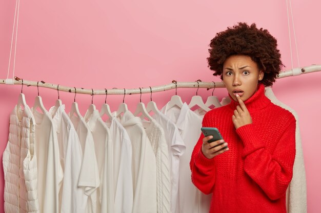 Shopping, tecnologia e concetto di persone. Lo stilista femminile riccio nervoso posa vicino a molti vestiti bianchi sugli scaffali, indossa un maglione lavorato a maglia rosso, utilizza il telefono cellulare,