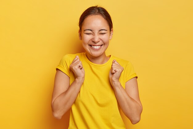 Sì, finalmente successo! Gioiosa ragazza coreana stringe i pugni con trionfo, chiude gli occhi dalla felicità e dalla gioia, ha un sorriso a trentadue denti, indossa abiti casual, isolato sul muro giallo, trionfa la vittoria