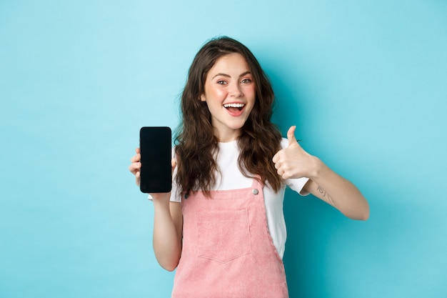 Sì, questo è buono. Sorridente ragazza carina che mostra pollice in su e schermo dello smartphone vuoto, consigliando l'applicazione o il negozio online, in piedi su sfondo blu.