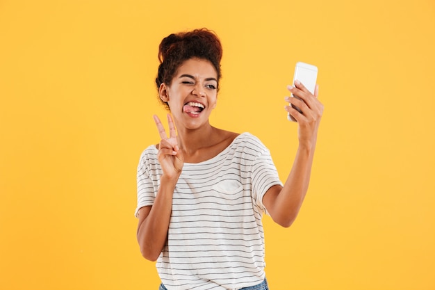 Signora africana divertente che fa smorfie e che fa selfie sul telefono isolato