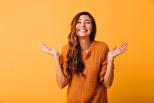 Signora alla moda con capelli marroni lunghi che ride divertente sul giallo. Modello femminile caucasico positivo in maglione di lana sorridente