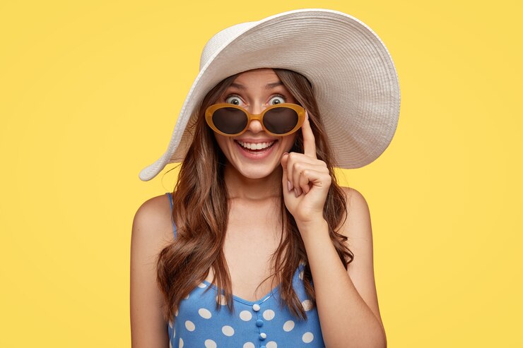 Foto gratuito signora alla moda con espressione felice, indossa cappello bianco e occhiali da sole, trova un hotel dove soggiornare durante le vacanze, pronta per andare in spiaggia, isolata sopra il muro giallo. turismo e concetto di ora legale