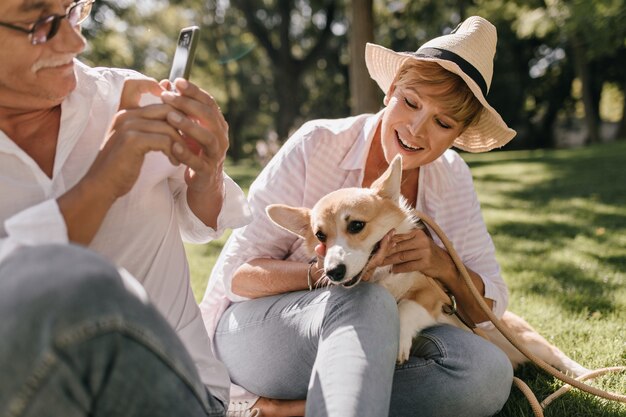 Signora alla moda con i capelli corti in cappello e camicia rosa sorridente, seduta sull'erba e in posa con corgi e uomo con lo smartphone nel parco.