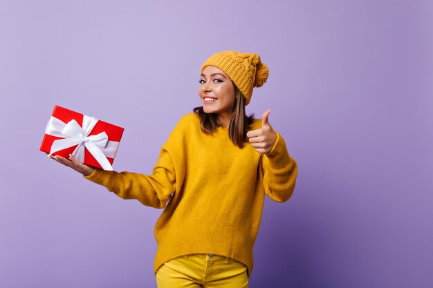 Signora allegra in cappello giallo alla moda ballando con regalo di compleanno. Ragazza caucasica di risata che tiene il regalo del nuovo anno sulla porpora.