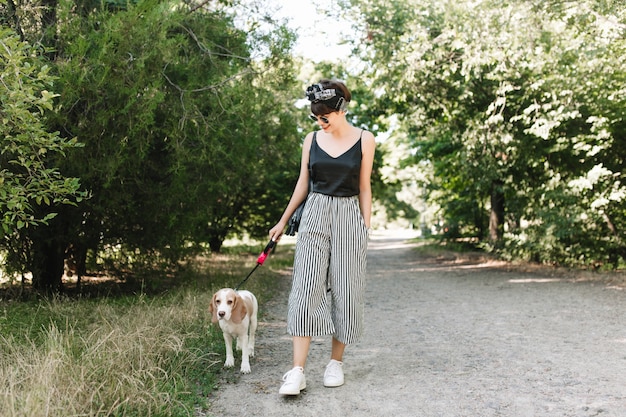 Signora allegra in scarpe da ginnastica bianche che cammina con il cane beagle nel parco in una giornata di sole, godendo del bel tempo