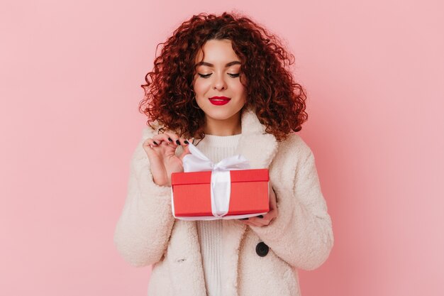 Signora attraente che tiene scatola regalo rossa con nastro bianco su spazio rosa. Istantanea di una ragazza riccia con un vestito di lana leggera.