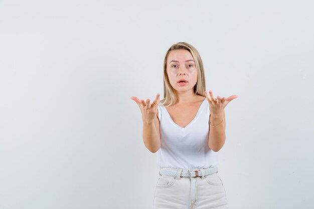 Signora bionda che allunga le mani nel gesto interrogativo in singoletto, pantaloni, vista frontale.