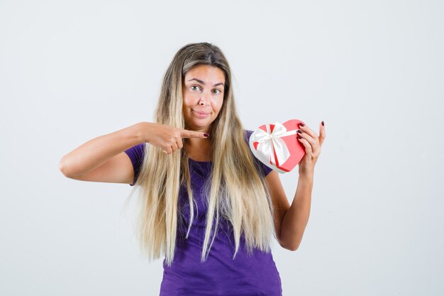 Signora bionda che indica al contenitore di regalo in maglietta viola e che sembra felice, vista frontale.