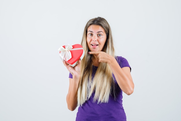 Signora bionda che indica al contenitore di regalo in maglietta viola e sembra allegra, vista frontale.