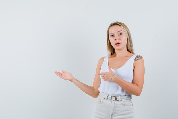 Signora bionda che indica da parte, mostrando qualcosa in singoletto, pantaloni e guardando fiducioso, vista frontale.