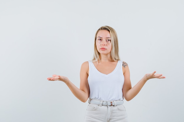 Signora bionda che mostra gesto impotente in canottiera, pantaloni e sguardo confuso