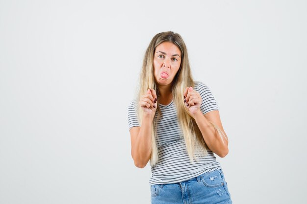 Signora bionda che mostra il gesto del fico, che attacca fuori la lingua in maglietta a righe, jeans e che sembra dispettosa. vista frontale.