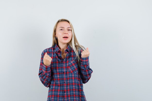 Signora bionda che mostra il gesto del vincitore in camicia casual e sembra fortunato, vista frontale.