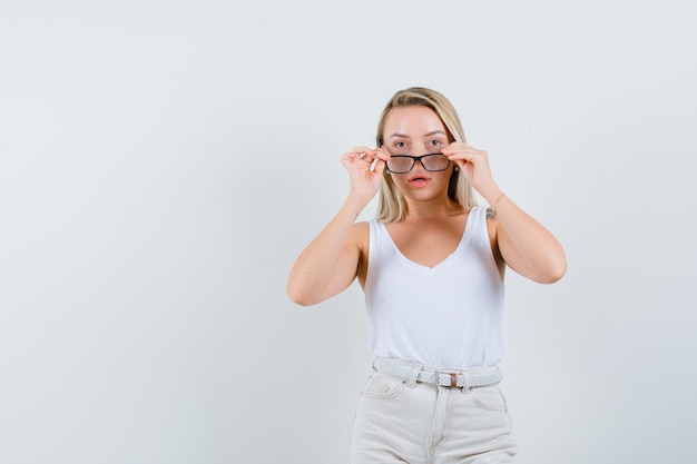 Signora bionda che osserva sopra gli occhiali in canottiera, pantaloni e guardando sorpreso, vista frontale.