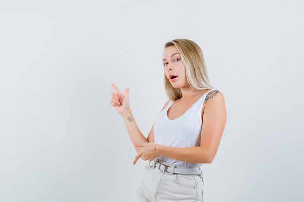 Signora bionda che punta le dita su e giù in canottiera, pantaloni e sguardo esitante, vista frontale.