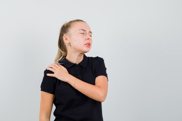 Signora bionda che soffre di dolore alla spalla in maglietta nera e sembra affaticata. vista frontale.