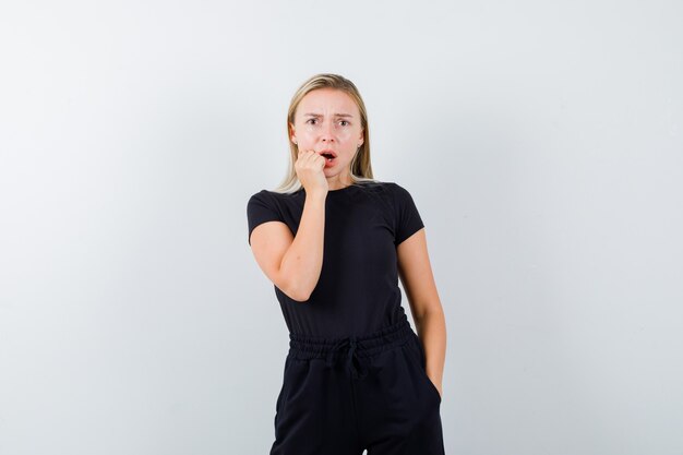 Signora bionda che tiene la mano vicino alla bocca aperta in vestito nero e che sembra ansiosa, vista frontale.